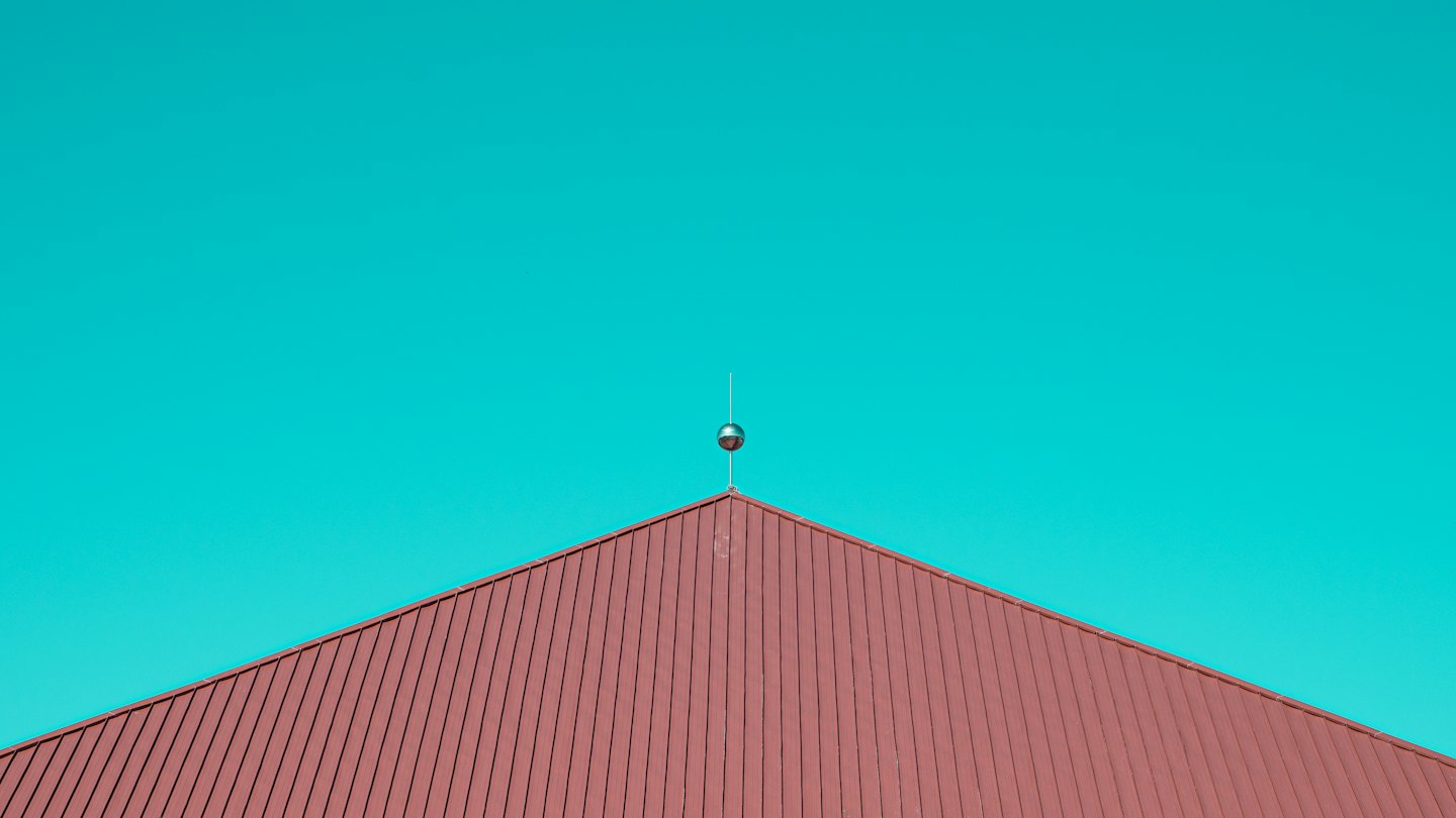 Red metal roof peak with lightning rod against turquoise sky.